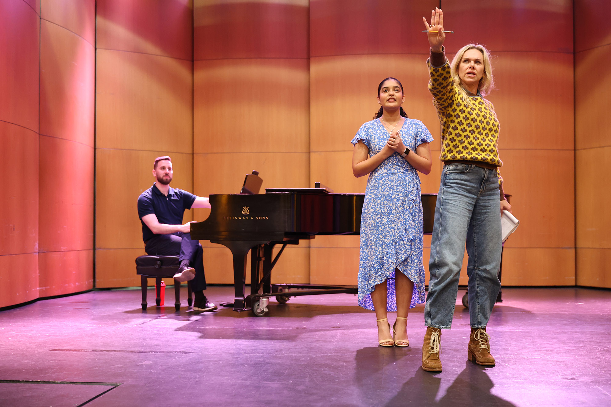 Broadway star Laura Bell Bundy gestures expressively while coaching a UNCP Musical Theatre student on stage during the pre-show masterclass at the Givens Performing Arts Center, with a pianist at a Steinway grand piano in the background.