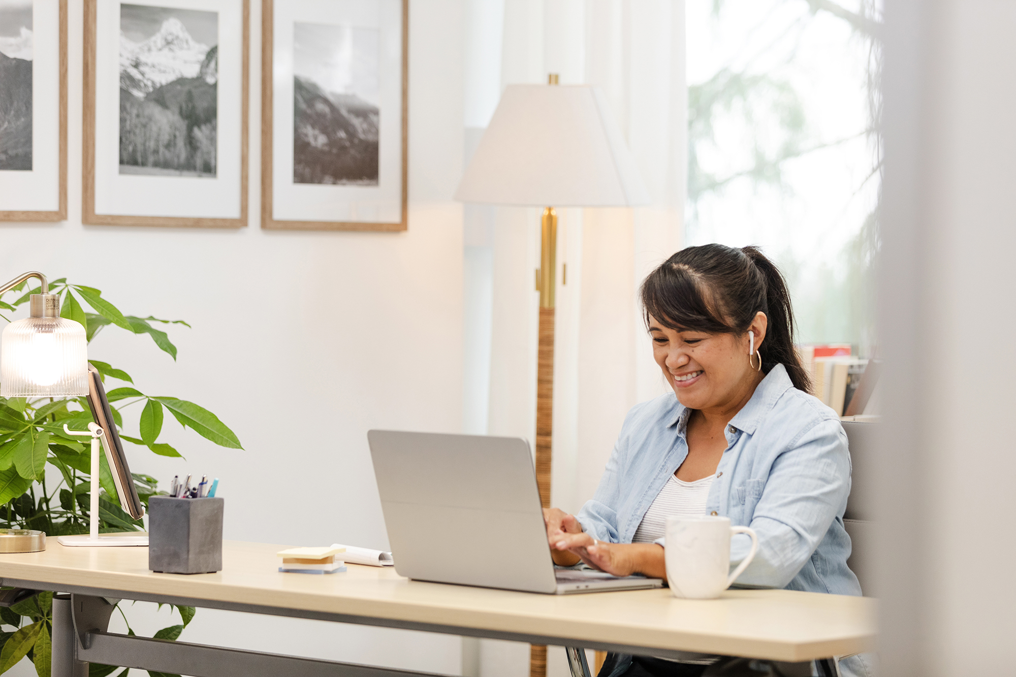 woman at table with laptop wearing earbuds and smiling with cup of coffee