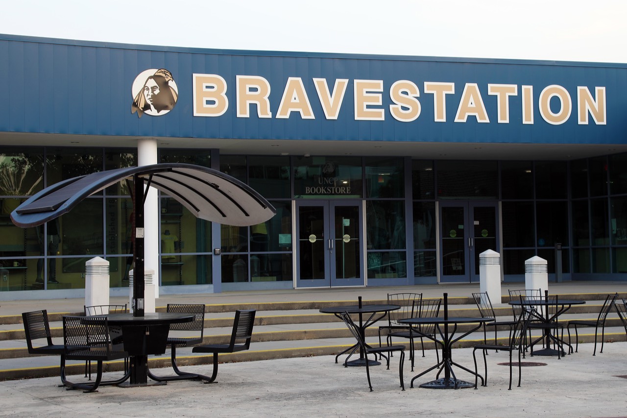 The BRAVESTATION at UNC Pembroke, featuring the entrance to the campus bookstore with large bold signage above the doors. In front, several tables and chairs are arranged for outdoor seating, including a solar-powered charging table with a curved canopy, providing students with a sustainable way to charge electronic devices while enjoying the space.