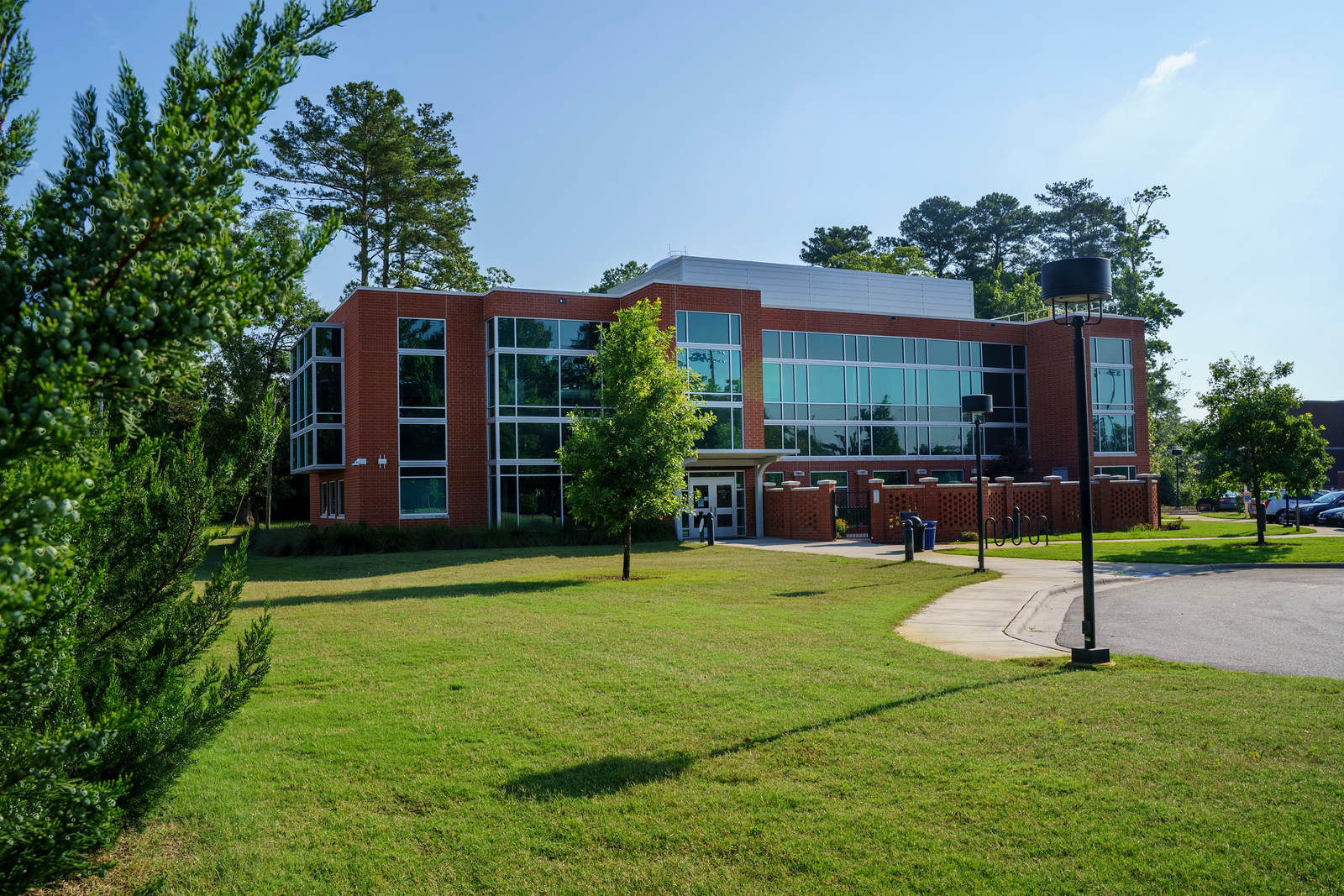 Brave Health building at UNC Pembroke, surrounded by green grass, trees, and a clear blue sky. The structure features large windows, bike racks, and shaded pathways, showcasing sustainable landscaping and architecture.