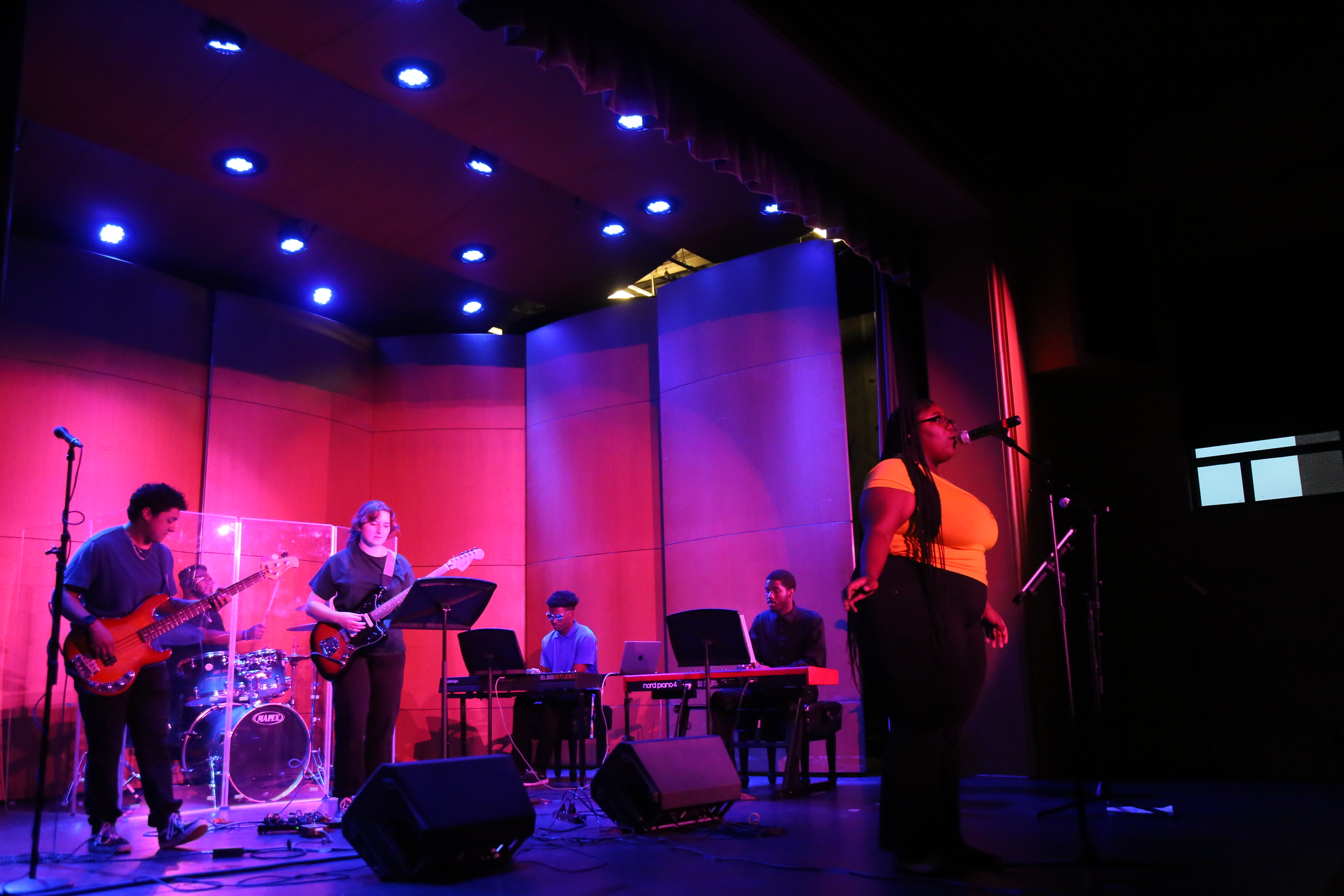 A student ensemble performs on stage under purple and blue lighting, with a singer in a yellow top standing at the microphone and musicians playing electric guitars, keyboards, and drums.