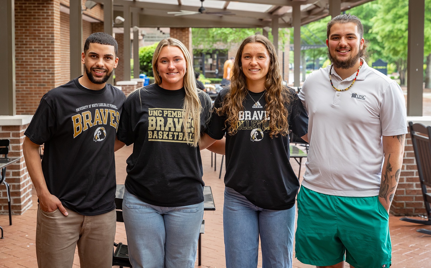 Four UNCP students smile together on campus wearing Braves and UNC Pembroke branded apparel.