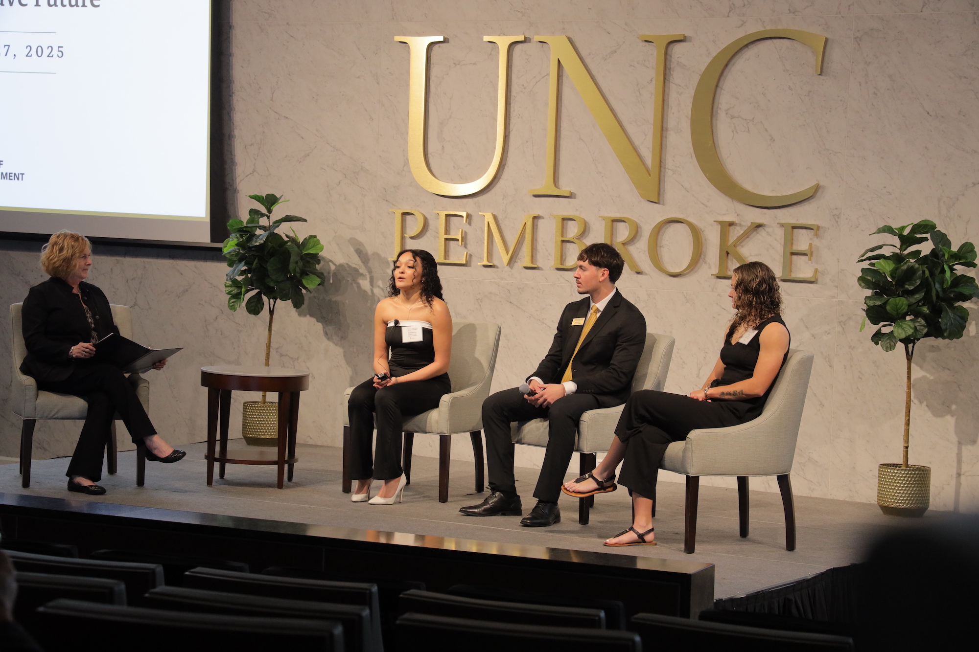 The VC of Advancement seats on a stage with 3 UNCP students.