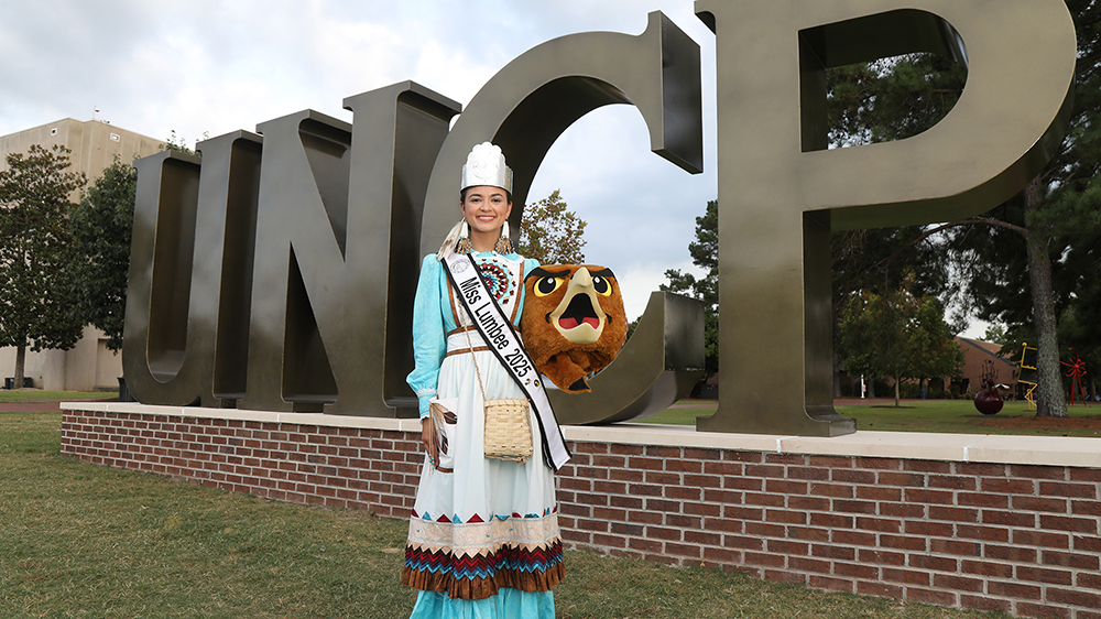 Miss Lumbee Taylor Davis stands in front of the UNCP letters while holding the head of the mascot, Brave Hawk