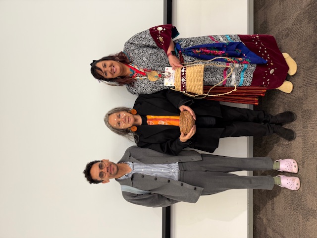 Three people stand together in a conference room, with one person holding a woven basket and another wearing traditional Indigenous-style regalia and holding a second basket.