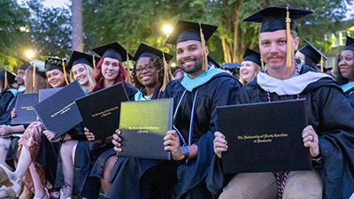 UNC Pembroke graduate students sit together outdoors during commencement, smiling and holding their diploma covers.