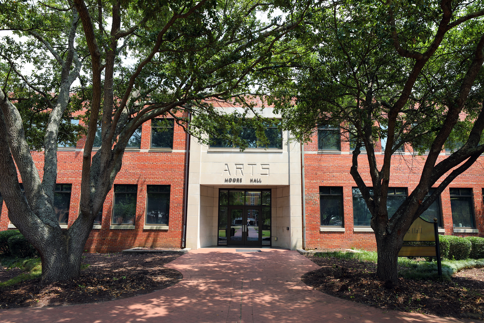 Front entrance of Moore Hall, home to the Department of Music and Arts at UNC Pembroke, framed by large trees and a brick walkway on a sunny day.