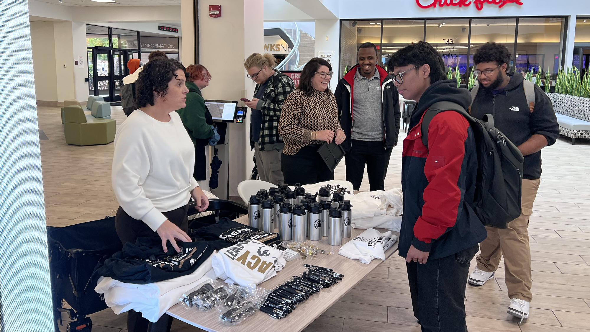 A fenale adminstaror stands at a table displaying tshirts and cups and is talking to several male college students wearing backpacks.