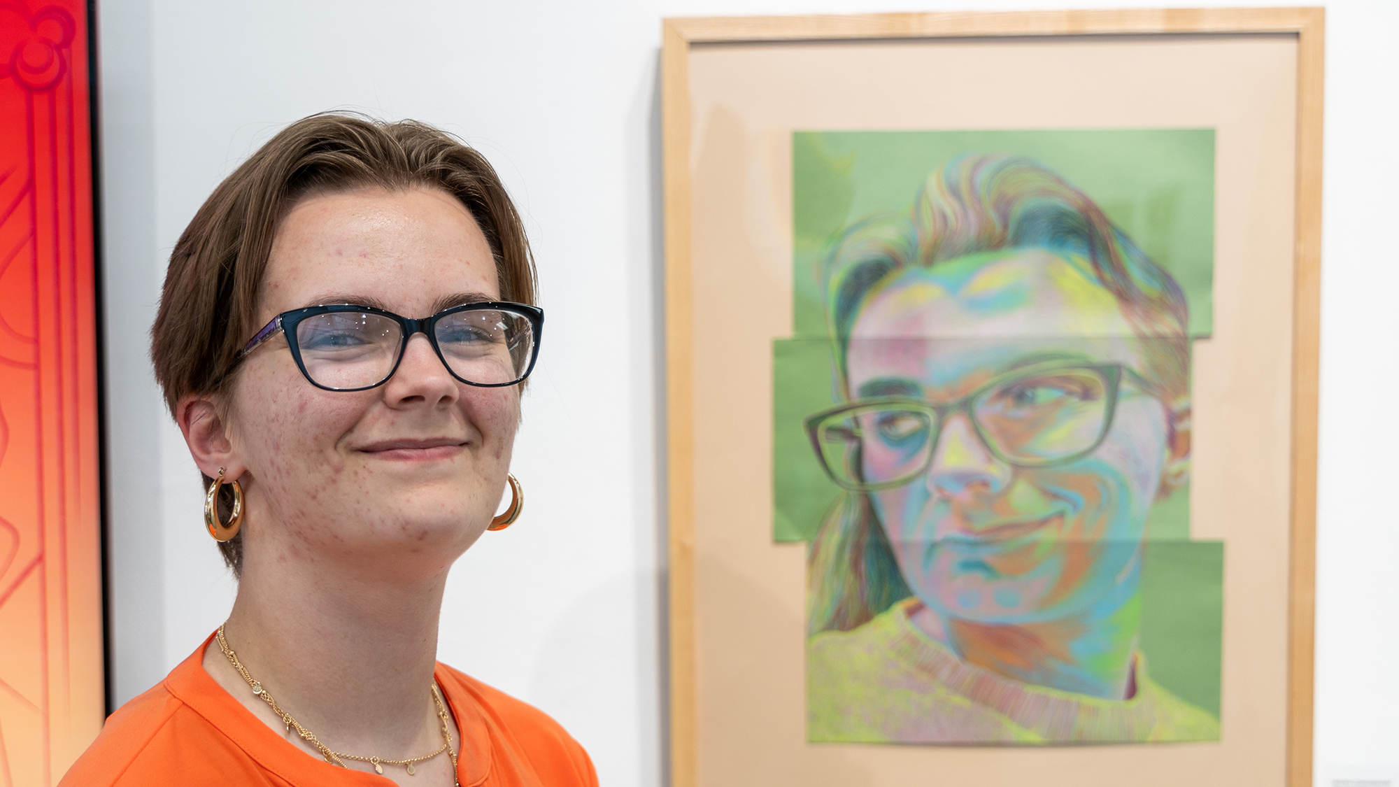 student smiling with self-portrait painting hanging on the wall to her left