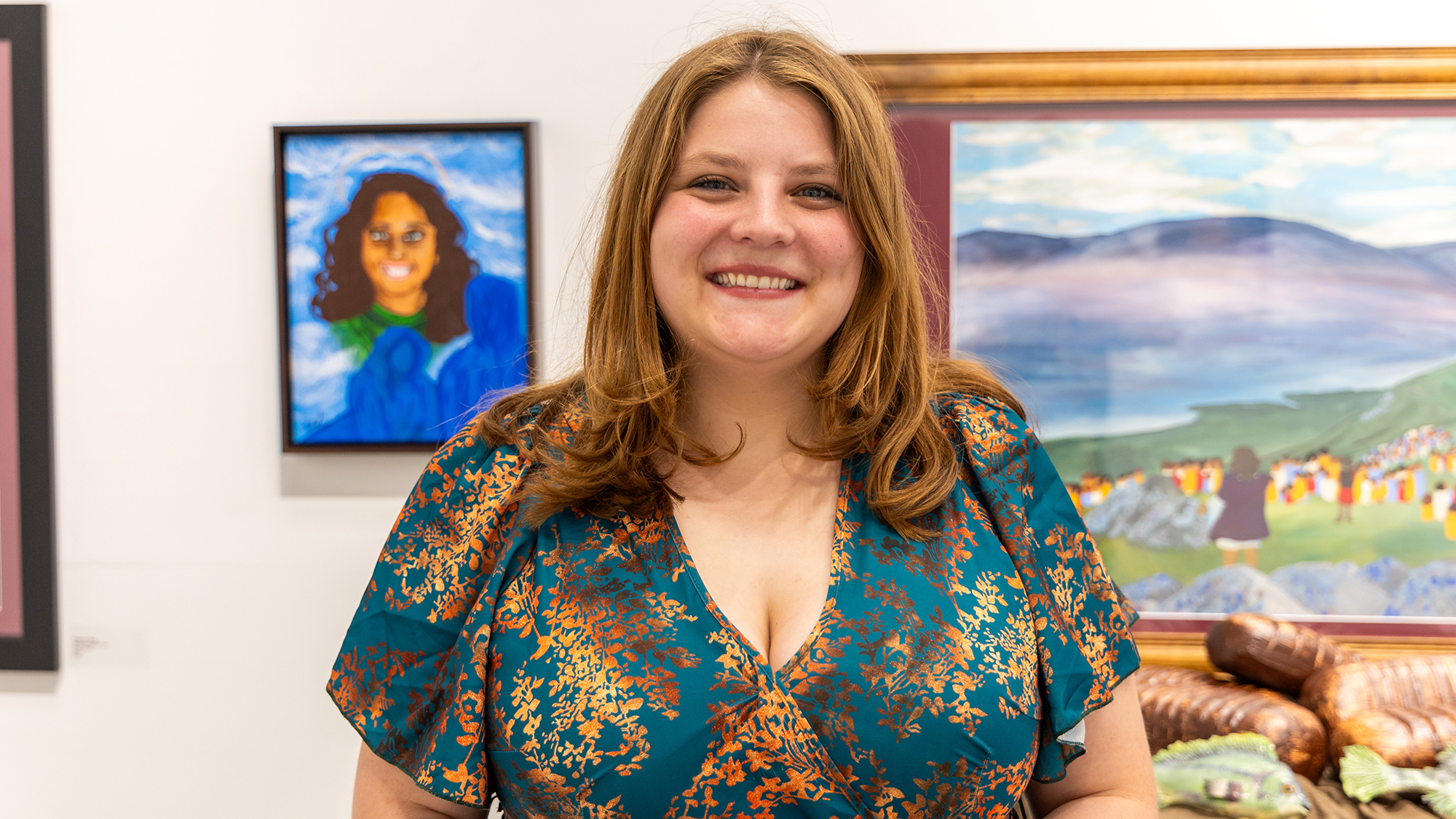student standing in front of paintings hanging on the wall - portrait over her right shoulder and a landscape over her right