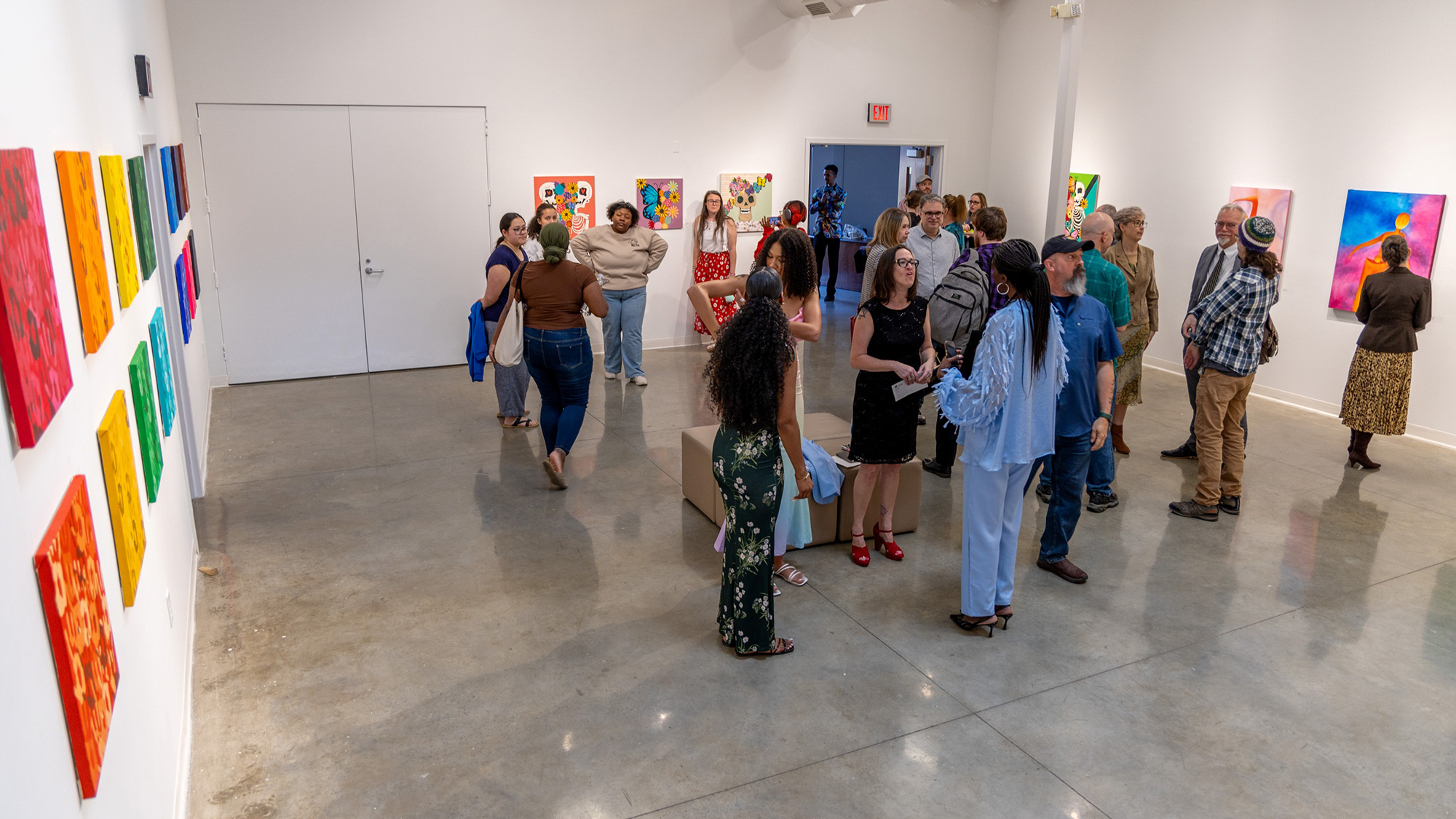 Art gallery reception full of people talking and looking at art mounted on white walls. Colorful paintings line the left wall shown in the image.