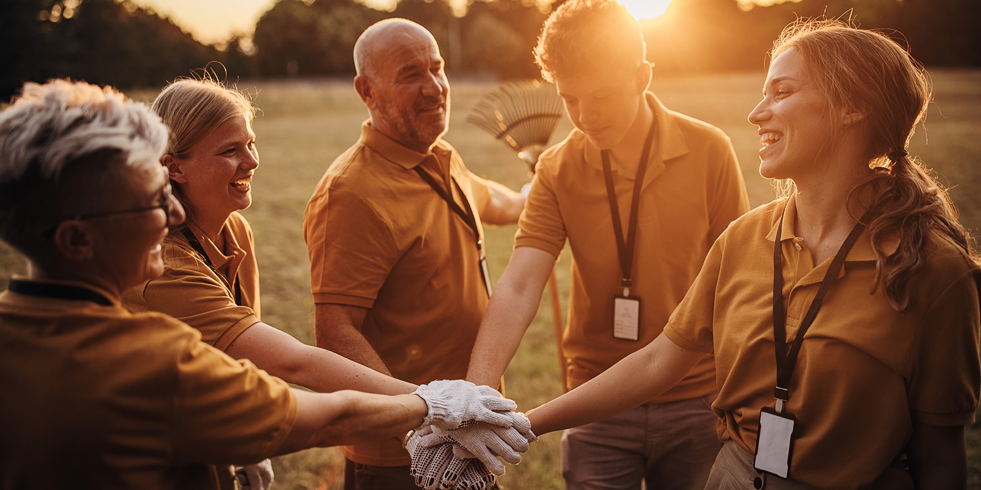 Stock image of a circle of people with their hands together int the center of the cirlce with the sun setting behind them.