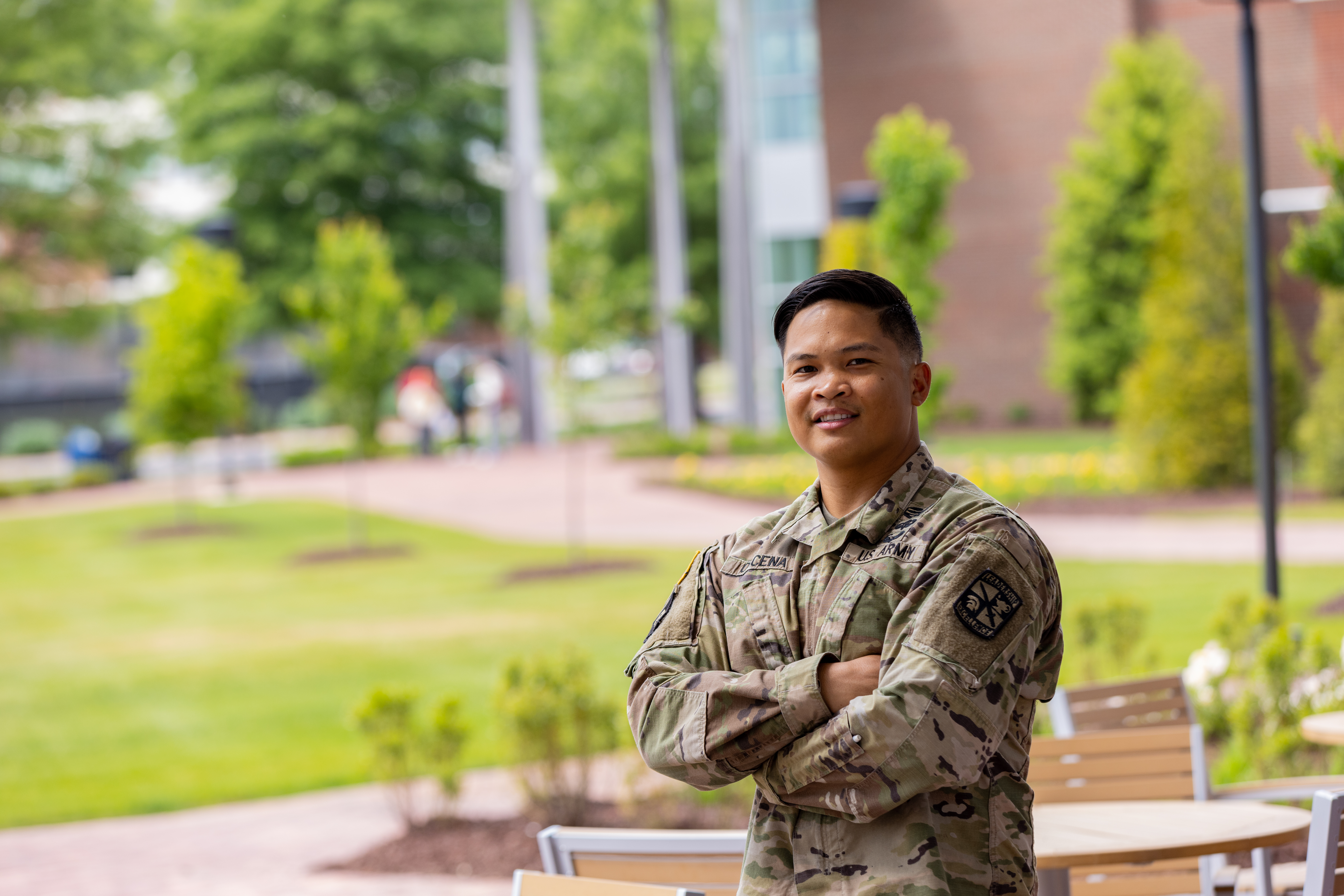 A person in a U.S. Army uniform standing outdoors with greenery and outdoor furniture in the background.