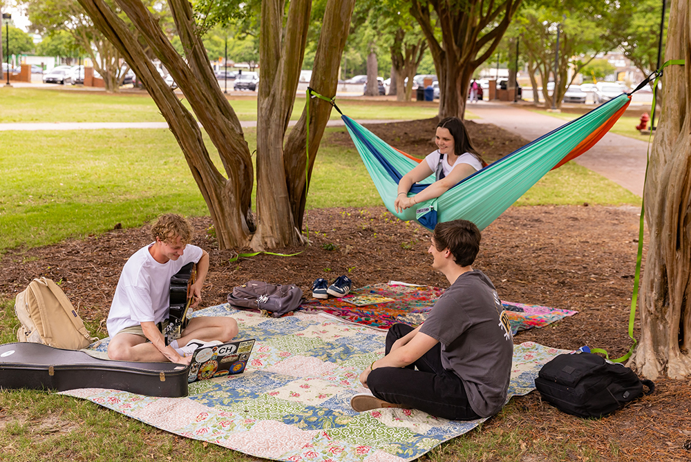 two students sitting on a blanket another in a hammock on campus as one of the students plays a guitar