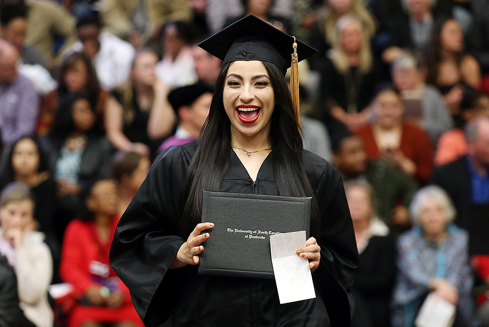 UNC Pembroke graduate in her black and goldcap and gown, holding up her diploma and smiling to the camera.