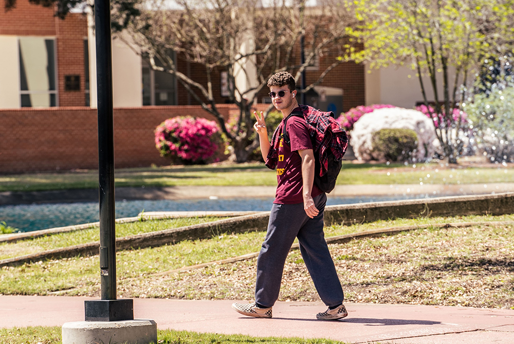 Student walking across UNC Pembroke campus. Bushes are in full bloom. The student looks into the camera and waves the two-finger peace sign.