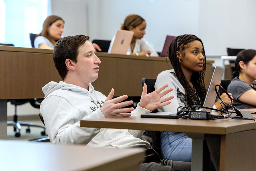 White male student at a classroom table next to a black female students, gesturing with his hands while talking to a professor, out of frame. This is a business class in UNC Pembroke's College of Business and Economics.