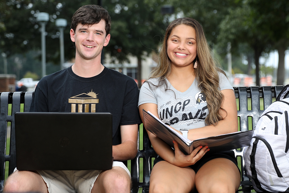 UNCP male and female students sitting on a bench with a computer