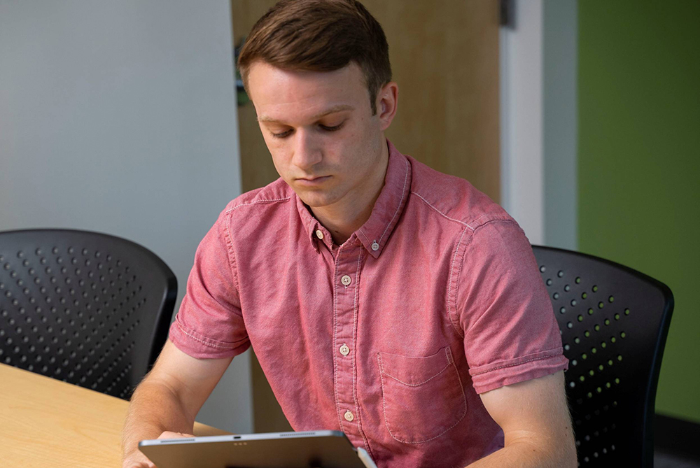 Young male adult holding a tablet while seated at a table.