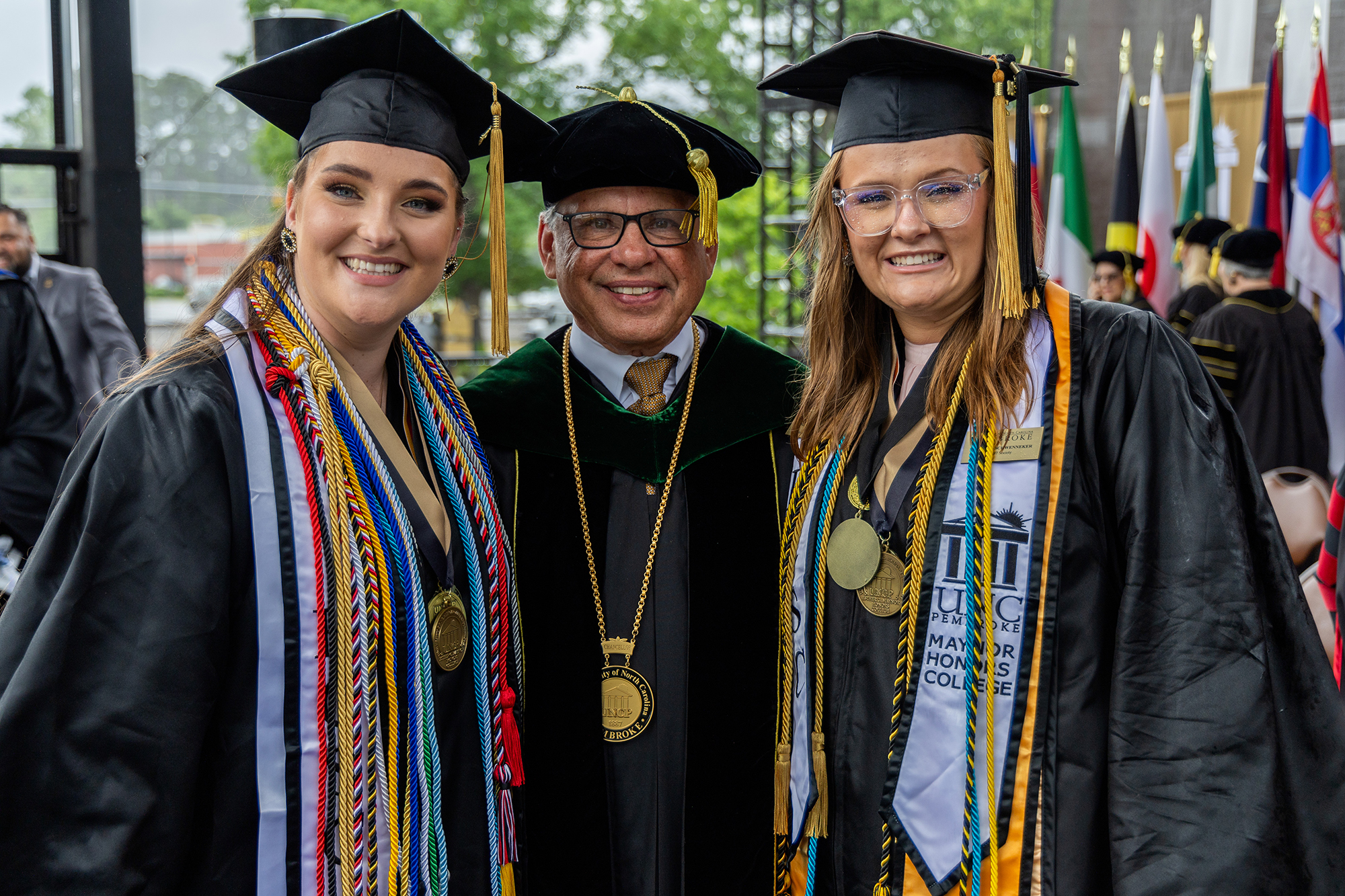 Aspen Andersson in cap and gown next to UNCP Chancellor Robin Gary Cummings and fellow graduate Madison Schwenneker