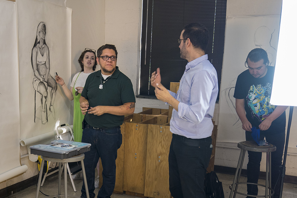 Art instructor talking with two students in a figure drawing class. The students are working in charcoal on large papers taped to the wall