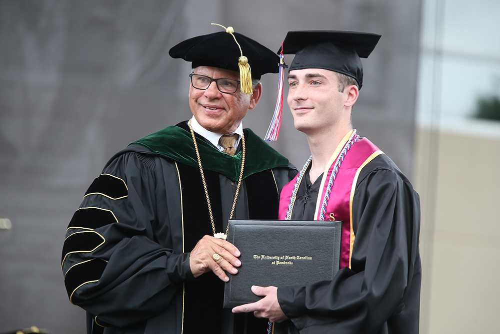 male graduate in cap and gown standing with UNCP Chancellor Dr. Robin Gary Cummings