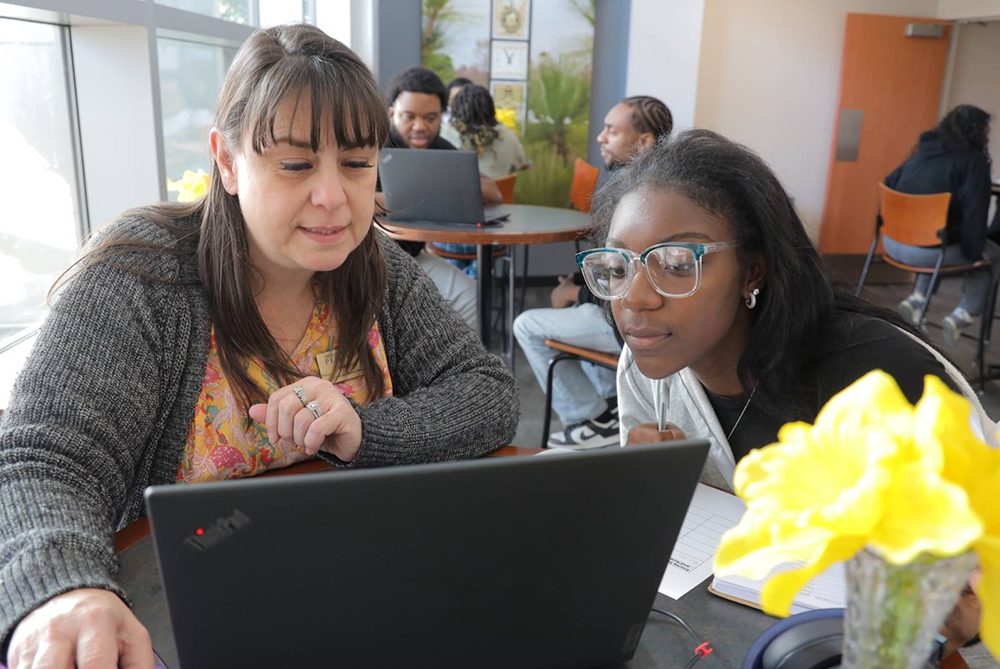 Advisor working student with advising on laptops in a modern, naturally lit room with large windows and indoor plants. Two individuals are in the foreground, one in a gray sweater and the other with long dark hair, with a yellow flower on the table in front of them.