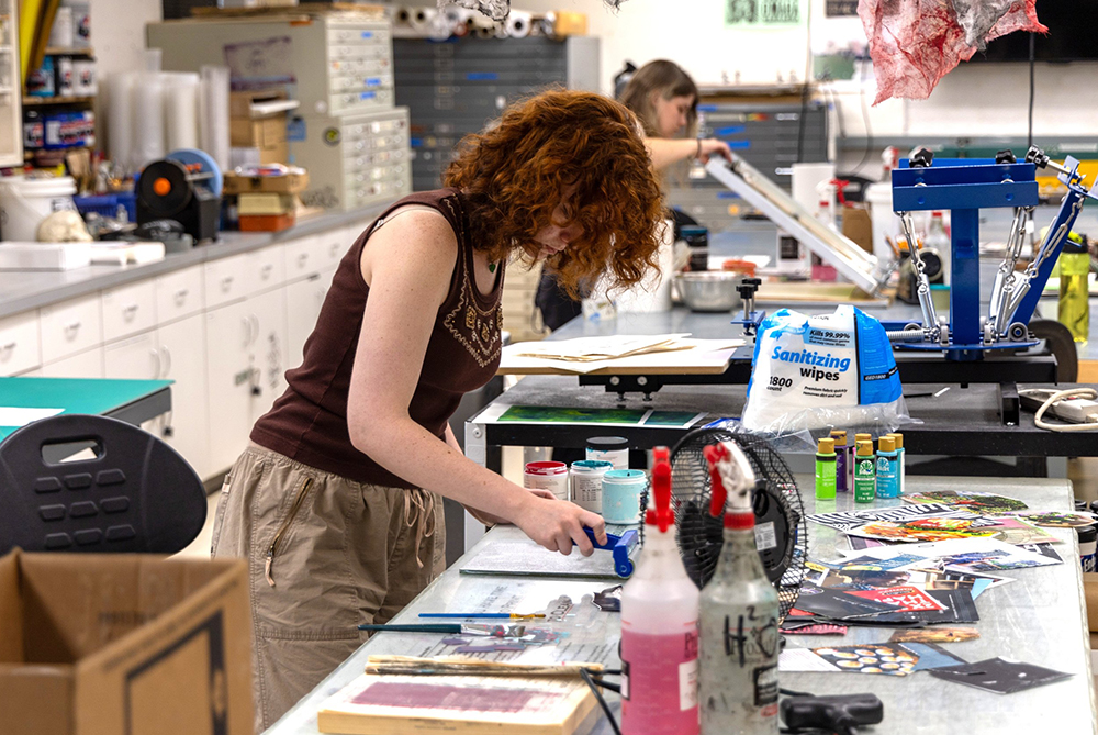 Two students in the printmaking studio at UNC Pembroke. The near student is rolling ink onto a woodcut. The far student is lifting a screen to change media for her screen printing.