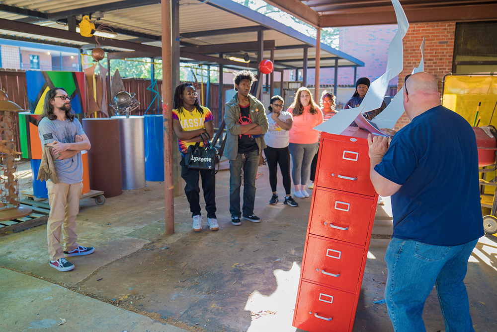 Professor speaking to the sculpture class in the outdoor portion of the sculpture studio amongs the students' large metal sculpture work at Locklear Hall, UNC Pembroke.