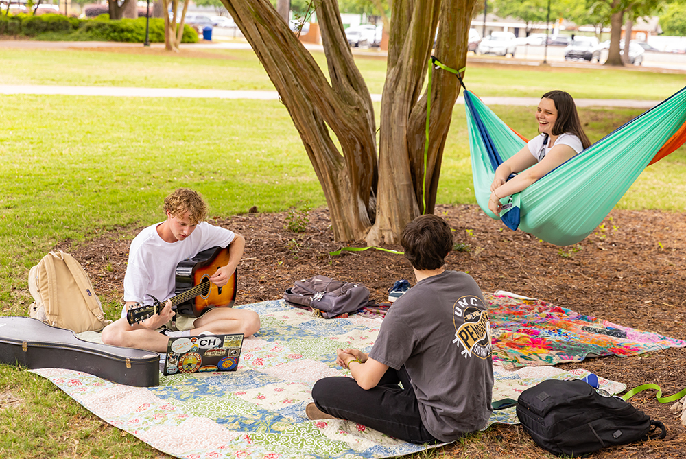 student sitting on blanket under tree playing guitar while another student sits with him and another is rocking in a hammock