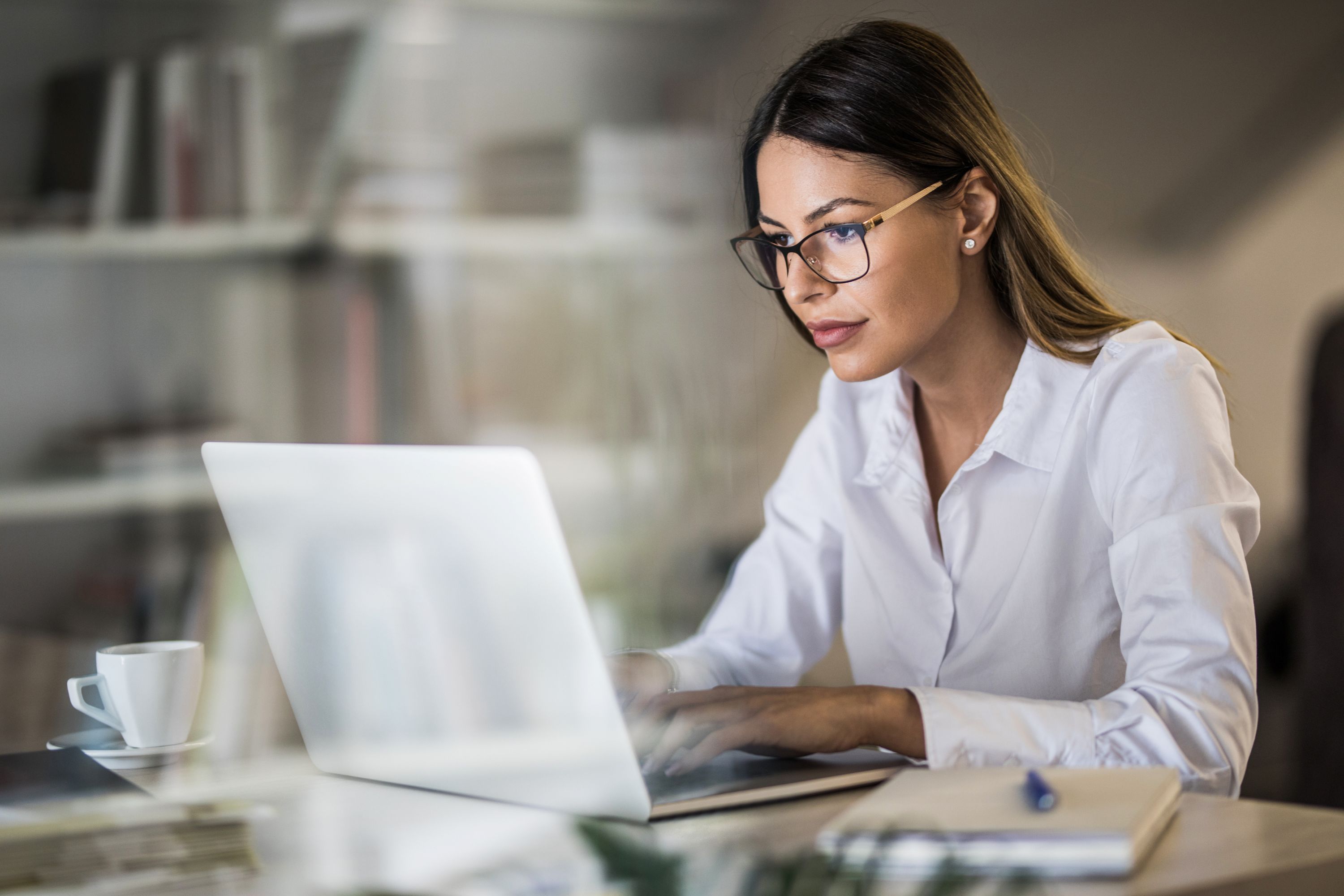 Woman with glasses in a button down shirt in a laptop with bookshelves behind her.