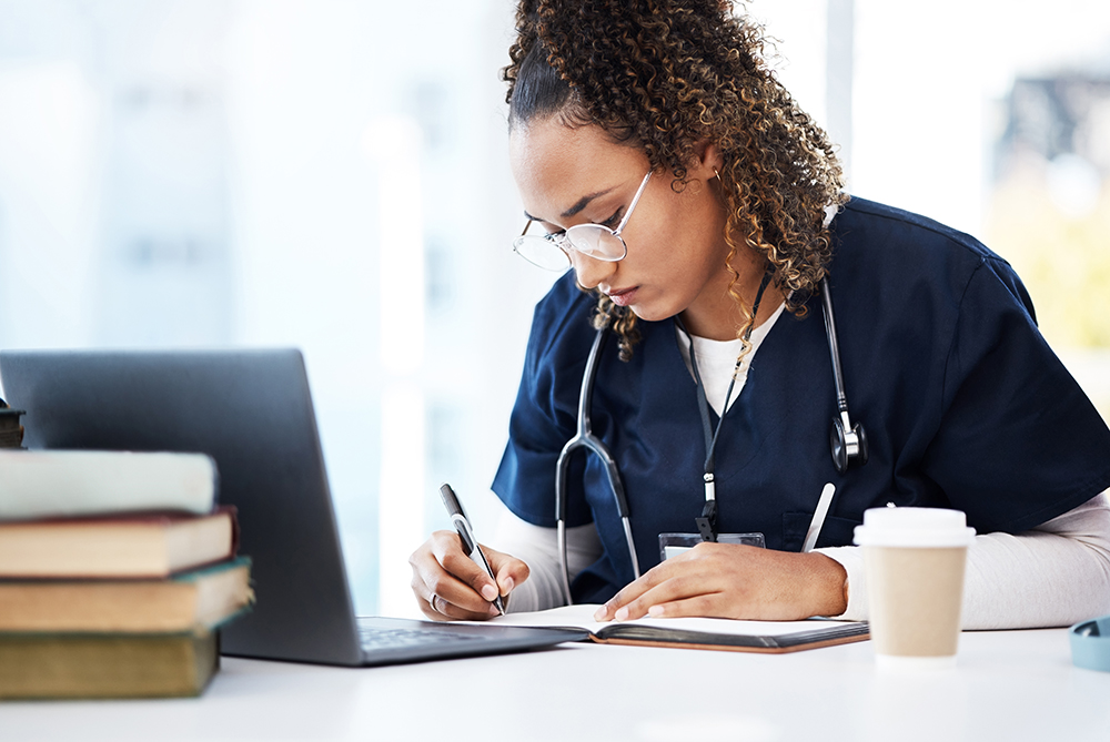 nurse with glasses looking down at laptop while surrounded by notebook as if she's studying