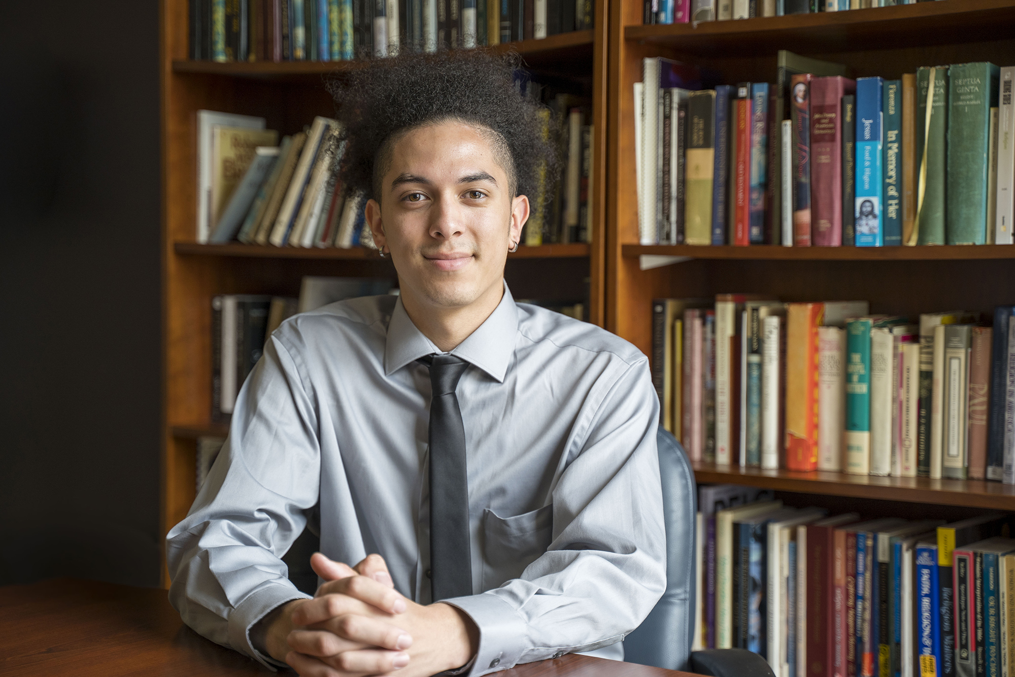 UNCP student Ahelayus Oxouzidis sitting in front of bookshelves at a table, smiling at the camera.