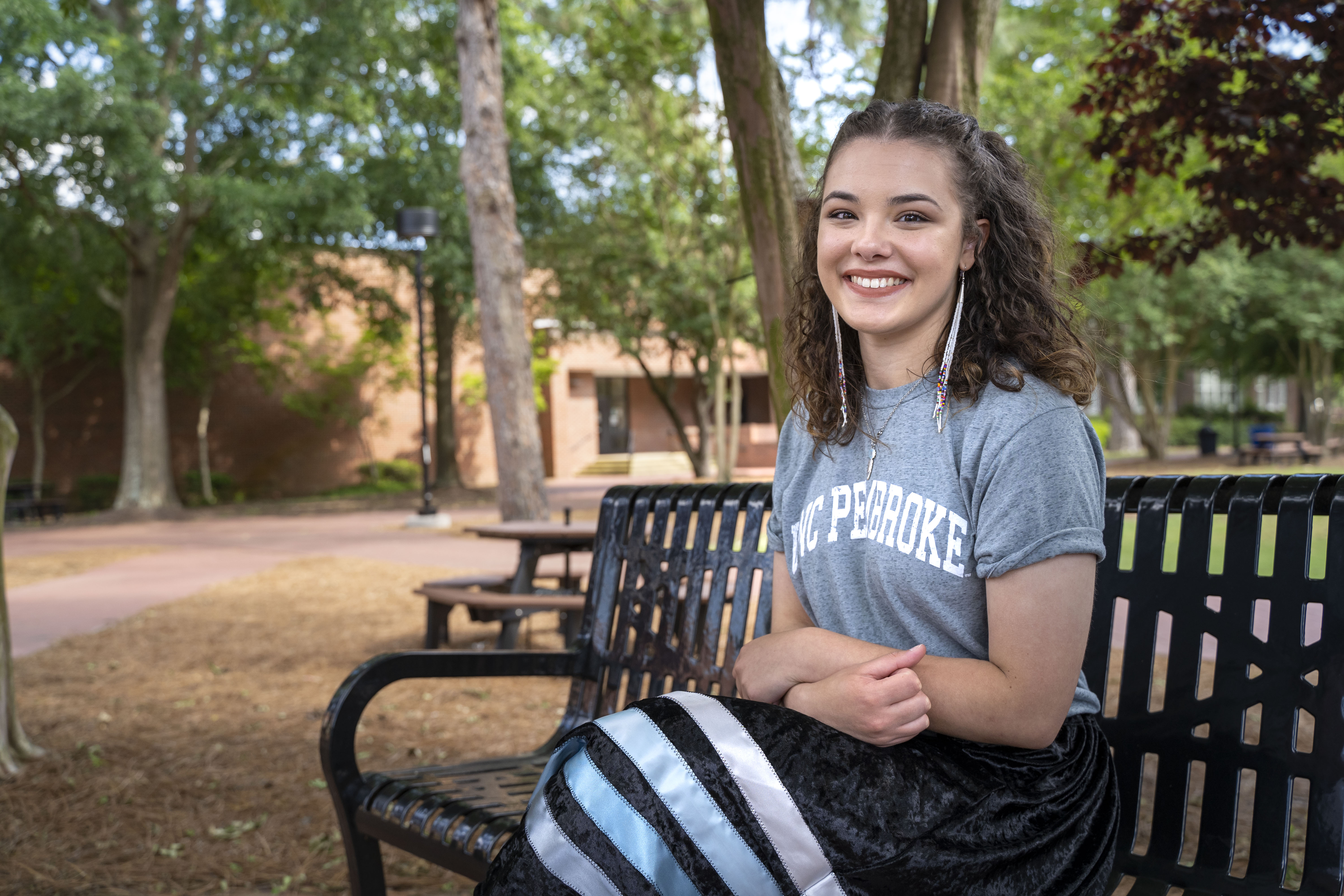 Group of people in UNCP undergraduates in caps and gowns, wearing honor cords and stoles. One face is blurred. Text and logos on the stoles are partially visible.