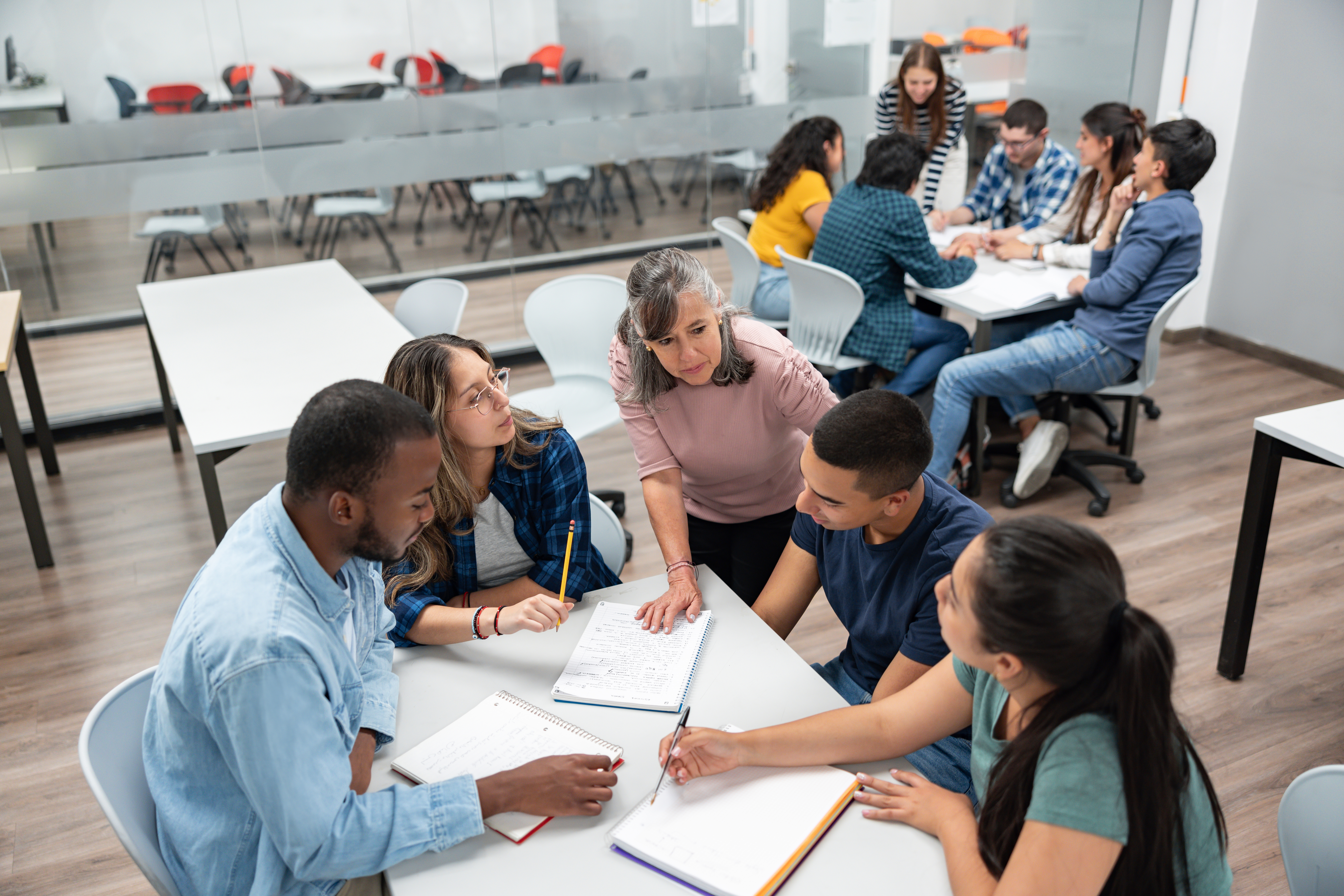 A group of diverse college students sits around a classroom table working on assignments, while a professor provides guidance. More students collaborate in the background.