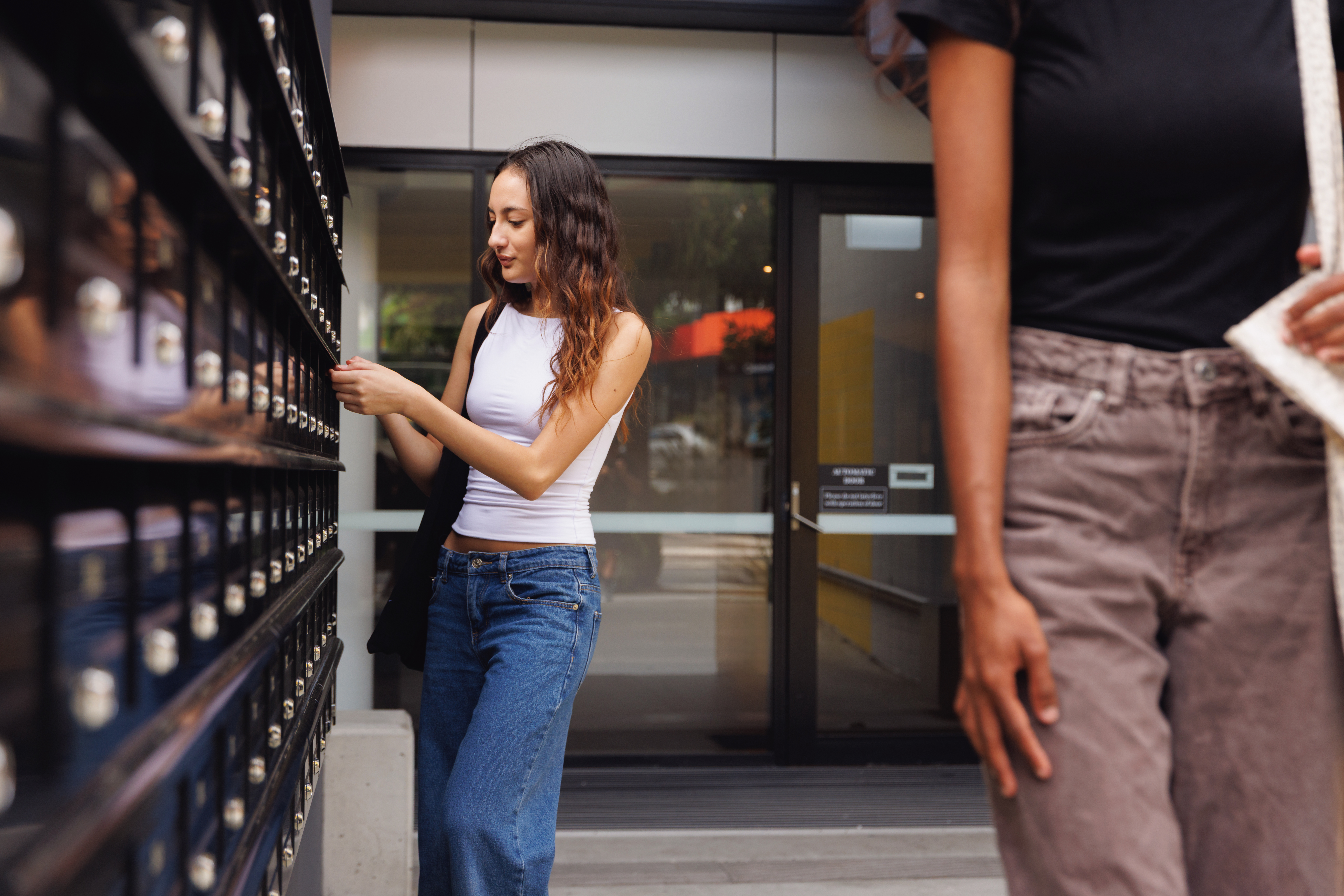 A student stands outside a campus building, opening a mailbox from a wall of student mailboxes. She is holding a key and wearing casual clothes, with another student walking by in the foreground.