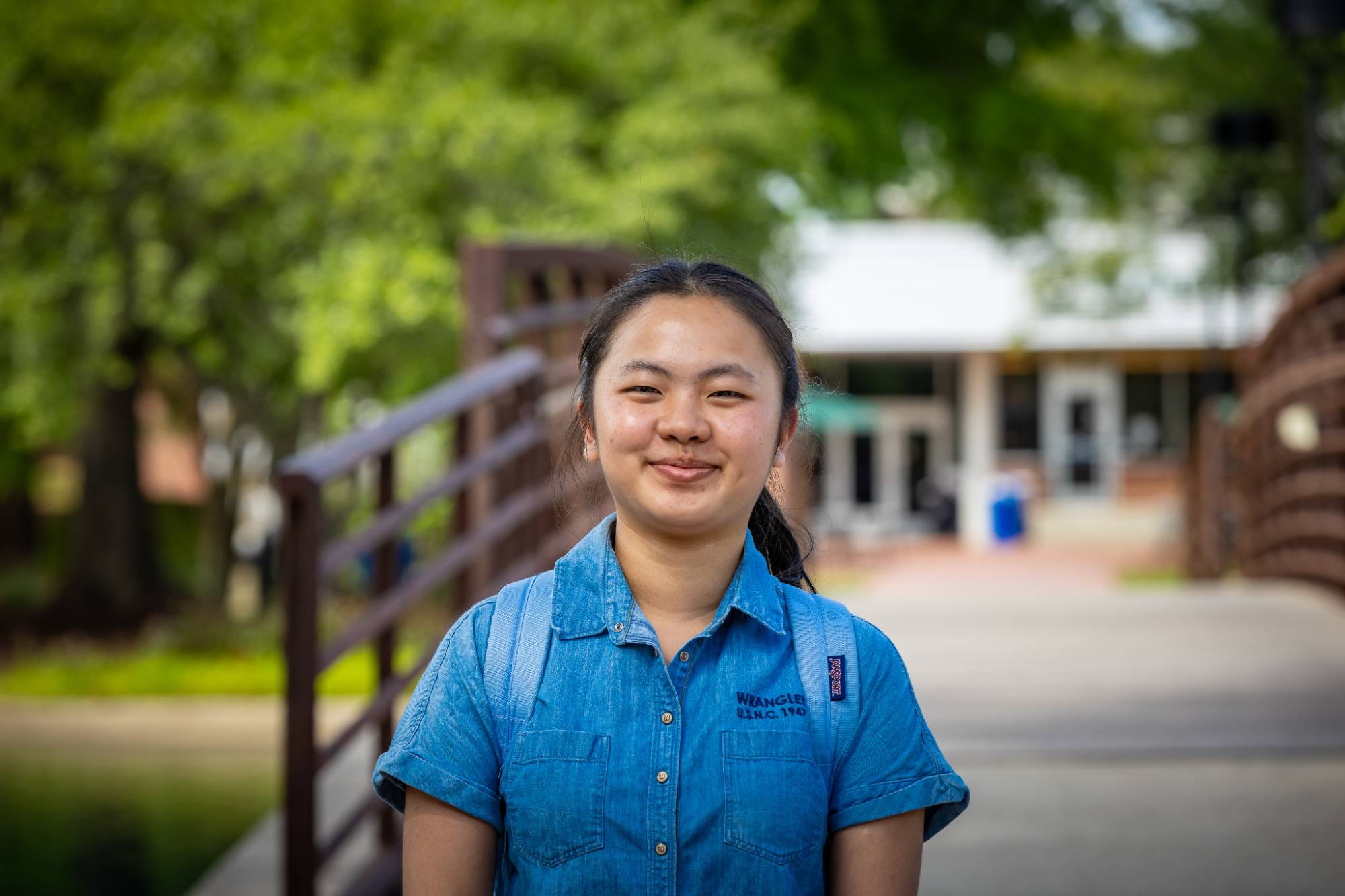 A smiling UNC Pembroke student stands on a pedestrian bridge on campus, wearing a blue denim shirt and backpack, with campus buildings and trees in the background