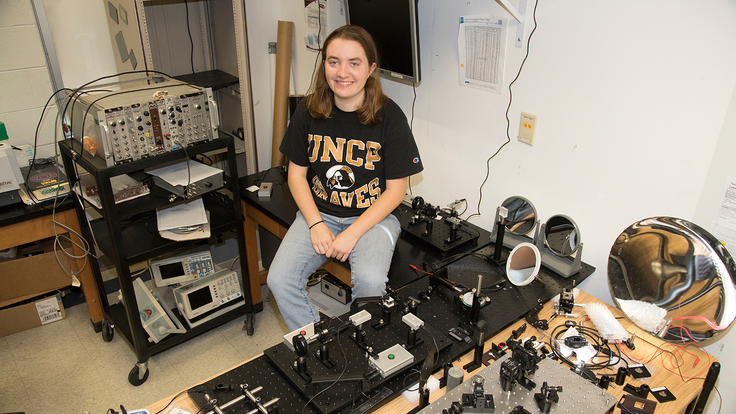 Female student wearing UNC Pembroke shirt sits in a lab surrounded by science equipment