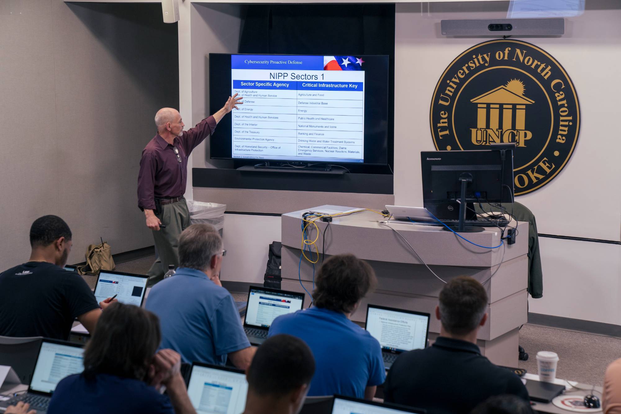 faculty standing at the front of a cybersecurity classroom at UNCP with computer screens visible