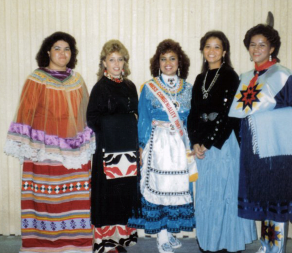 A group of American Indian women smile and pose in their tradtional regalia.