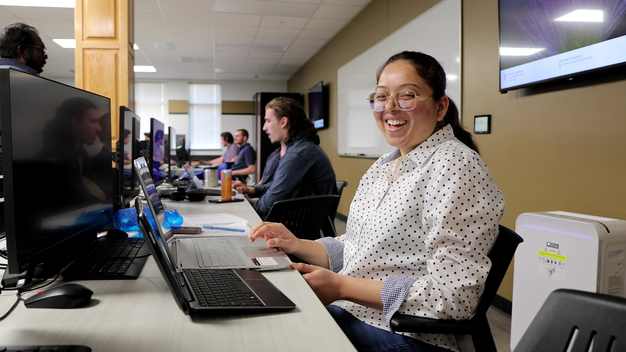 A UNCP Computer Science student smiles at the camera in a computer lab with two laptops open, with fellow students visible in the background.