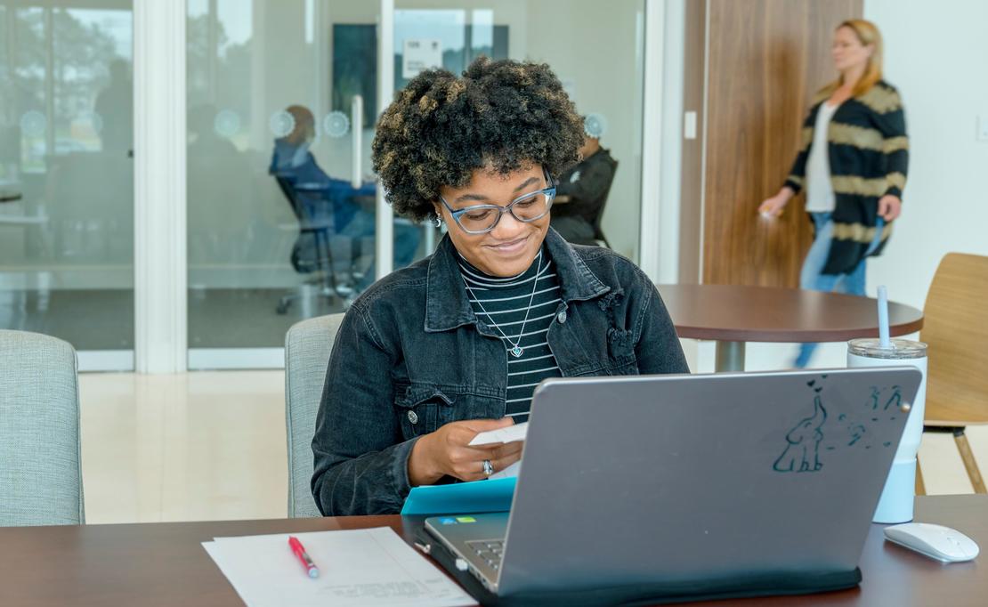 A photo of a student smiling while she is working on her computer in the James A. Thomas Hall building. 